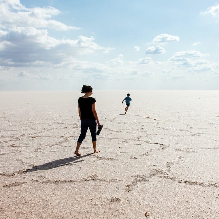 Auf dem Bild läuft eine junge Frau barfuß über einen weiten Sandstrand. Vor ihr rennt ein Kind, ebenfalls barfuß.