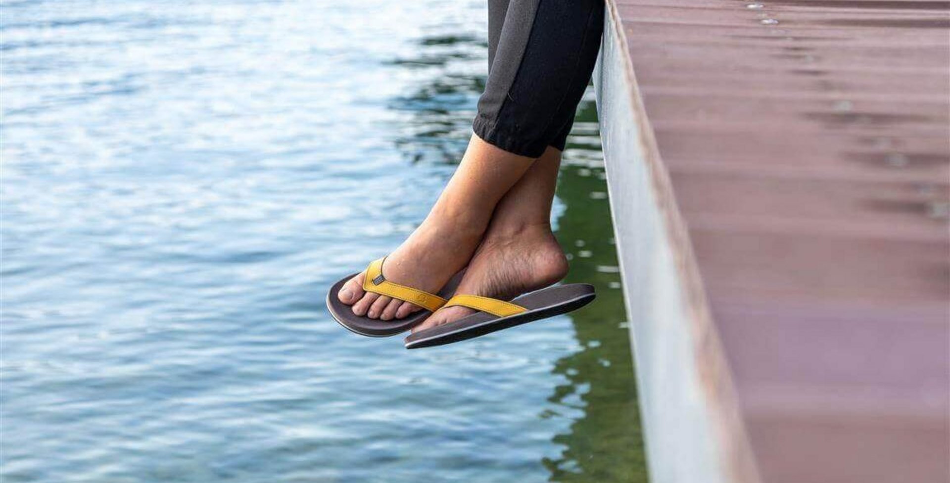 Bildausschnitt zweier Beine, die von einem Holzsteg über das Wasser eines Sees baumeln. An den Füßen trägt die Person ein Paar ErgoPad:ID Zehentrenner von Bauerfeind.